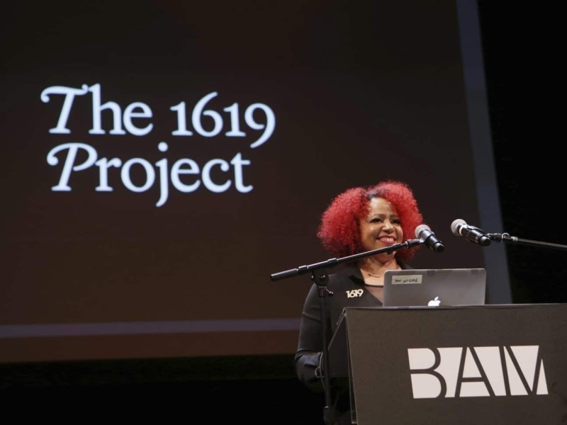 Nikole Hannah-Jones, a Black woman with red natural hair, presents The 1619 Project at a podium.