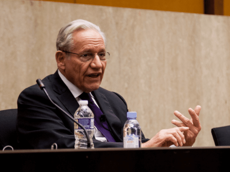 Bob Woodward, an elderly white man with glasses wearing a suit, in front of a speaker's mic.
