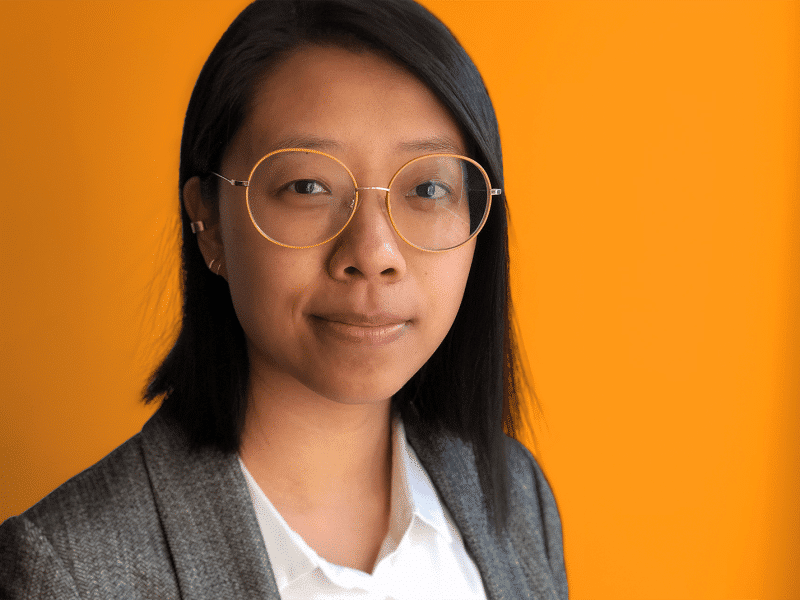 A headshot of Sisi Wei. She is in front of an orange background and wears a white collared shirt with a grey blazer.