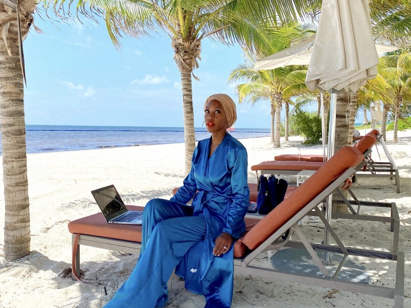 Imani Bashir sits sideways on an orange lounge chair on the beach. She wears a silk blue jumpsuit. Water and palm trees are in the background.