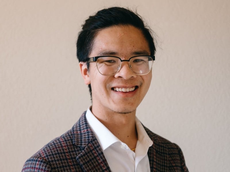 Headshot of Curtis Yee. He wears glasses, a white button-up collared shirt, and a plaid overcoat.
