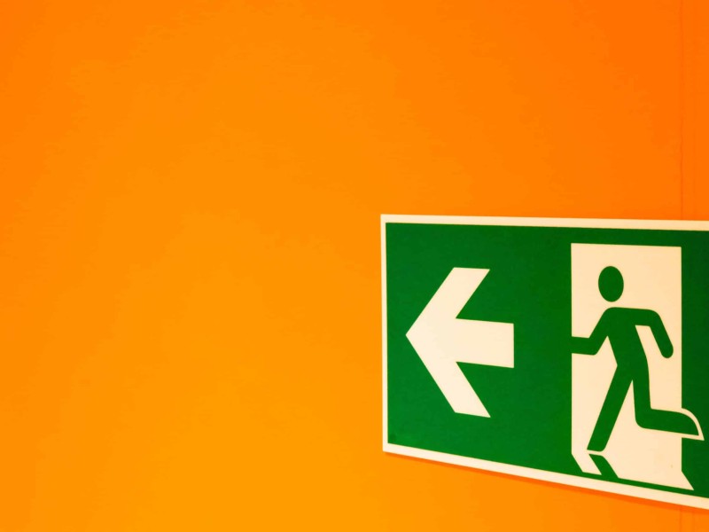 Green exit sign shows a running human figure and an arrow pointing left. The sign is right aligned on an orange background.