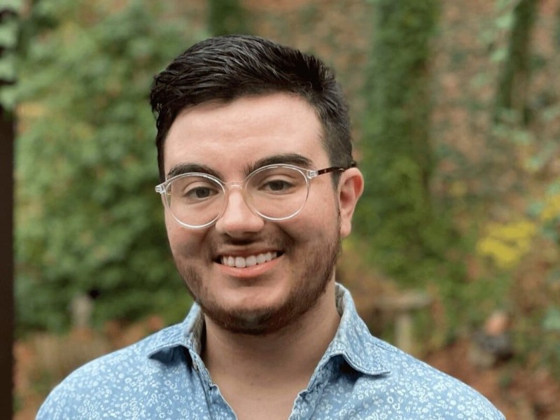 Headshot of Kam Burns. He has short brown hair, wears clear glasses frames, and a button-up collared shirt. Ivy grows on a wall in the background.