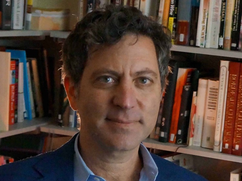 Headshot of Jamal Rayyis. He wear a blazer over a collared shirt and sits in front of a corner bookshelf.