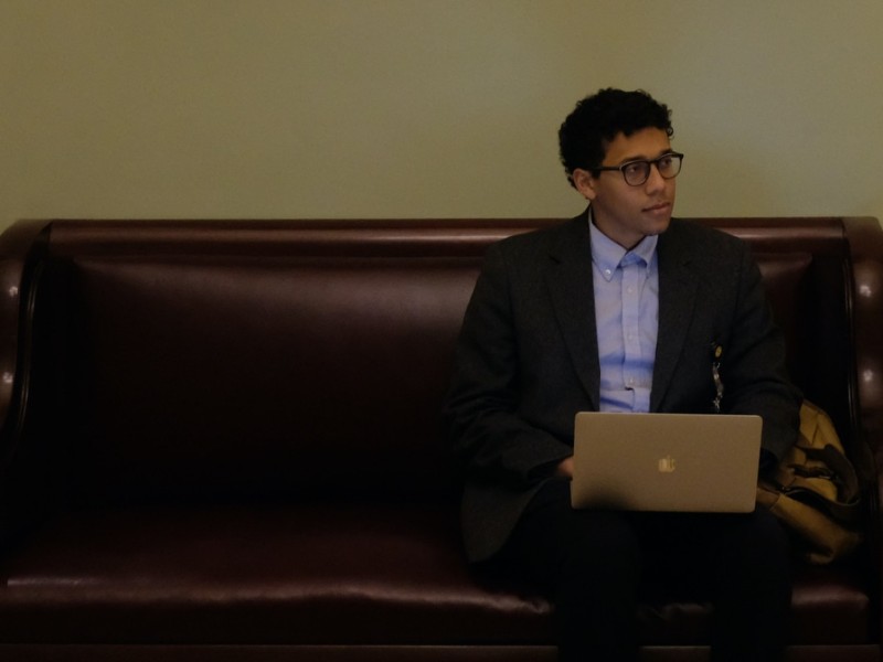 Gabe Schneider sits on a bench. He looks to the right of the frame and is holding a laptop in his lap. He wears glasses, a button up shirt, and a suit jacket.