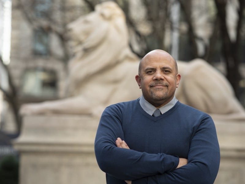 Dr. Thrasher stands in front of a lion sculpture with his arms crossed.
