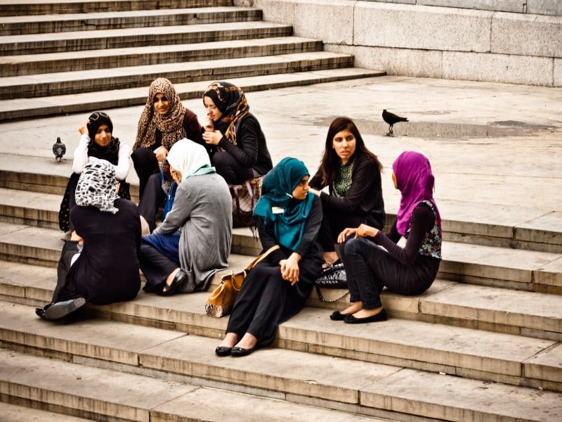 Young Muslim women, some hijabi and others not, sit on the steps of Trafalgar Square talking and laughing.