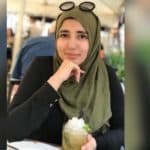 Anmol wears a long-sleeve plain black shirt and a green hijab with glasses atop her head. She smiles at the camera while holding a green tea.