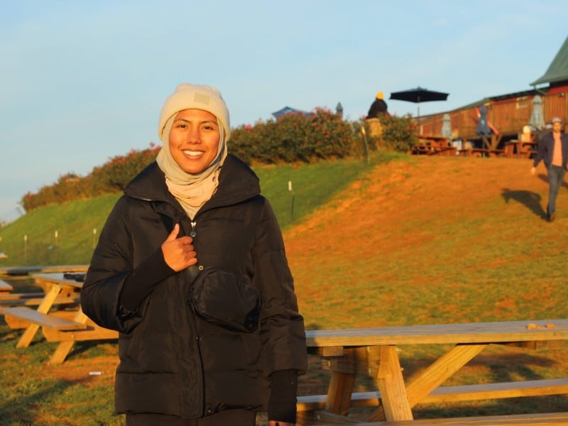 A headshot of Johana Bhuiyan. She has a thumb up and is in front of a grassy hill and picnic tables.