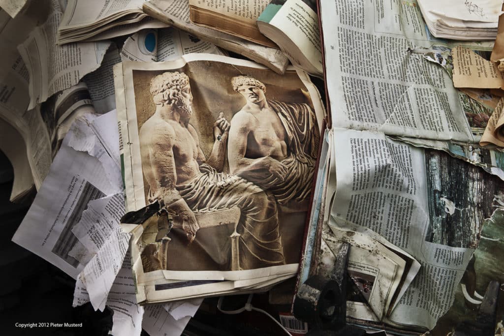 Crumpled newspapers lie atop each other. The image on the top newspaper is the focus, and features two human sculptures engaged in debate.