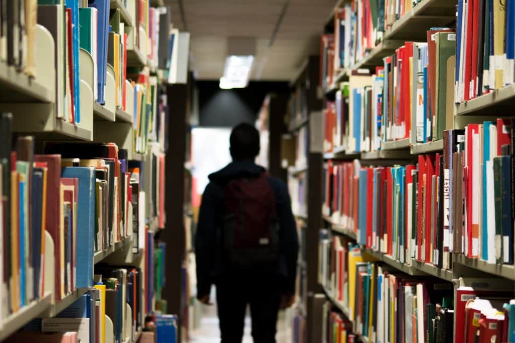 A high school student stands between two bookshelves in a library.