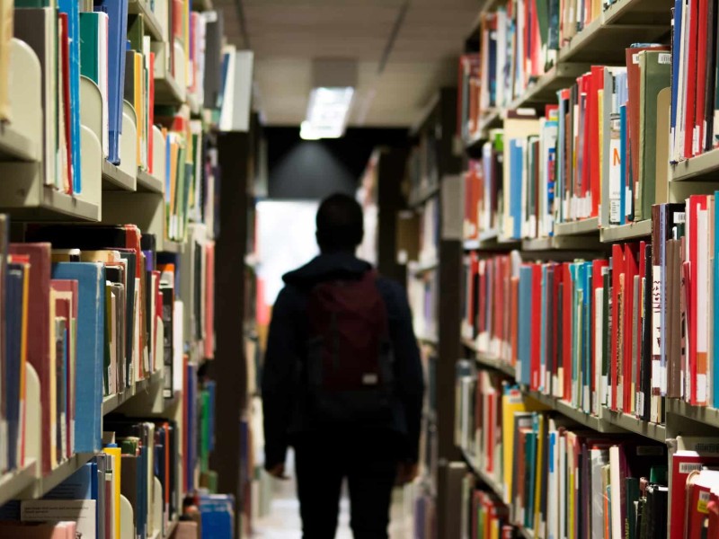 A high school student stands between two bookshelves in a library.