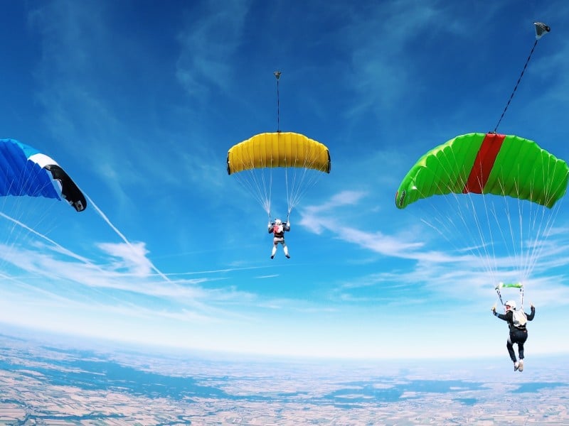 An image of three people parachuting over a landscape. The leftmost parachute is blue and black, the middle parachute is yellow, and the parachute on the right is green and red.