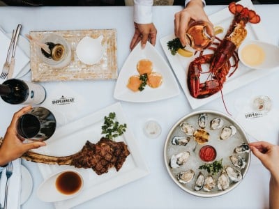 A flat lay of food. Napkins read "The Diplomat," the name of a now-closed Sacramento steakhouse. Among the platters are a dessert, lobster, oysters, and a tomahawk steak. Four diners' hands are visible in the photo. Two diners are holding glasses of wine, red and rosé specifically.