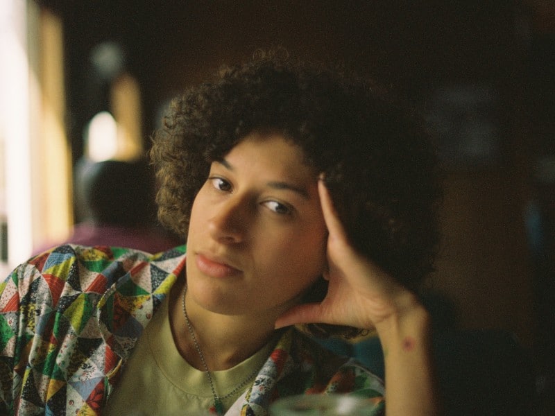 The author, Izzy Johnson, leans their head on their hand, looking at the camera with a neutral expression. They have an afro and are wearing a strawberry necklace, a short-sleeve rainbow patchwork square button-up and a pale green shirt.