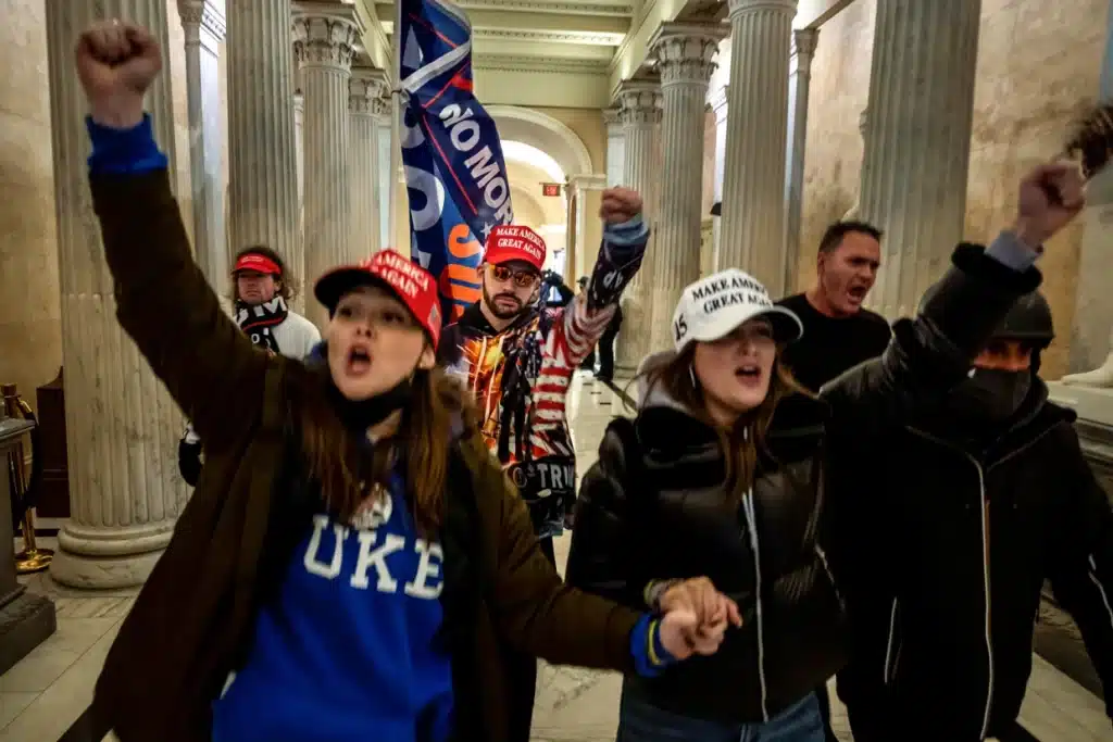 Two white women, holding hands, wear "Make America Great Again" hats and lead a group through the White House halls during the Jan. 6, 2021 insurrection. 