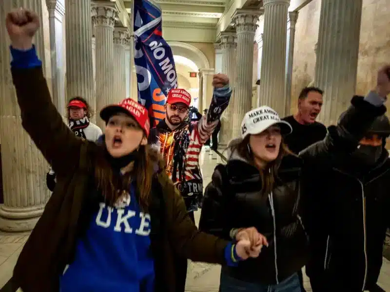 Two white women, holding hands, wear "Make America Great Again" hats and lead a group through the White House halls during the Jan. 6, 2021 insurrection.