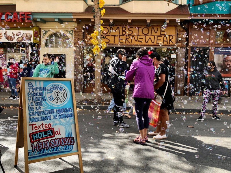 People gather in a street surrounded by a storm of bubbles for a Día de los Muertos celebration next to a Spanish sign from Bay Area news outlet El Timpano, encouraging attendees to sign up for their outlet. The sign reads "El Tímpano escucha y realza la voz de la comunídad Híspano hablante de Oakland. Textea HOLA al 15108008305."