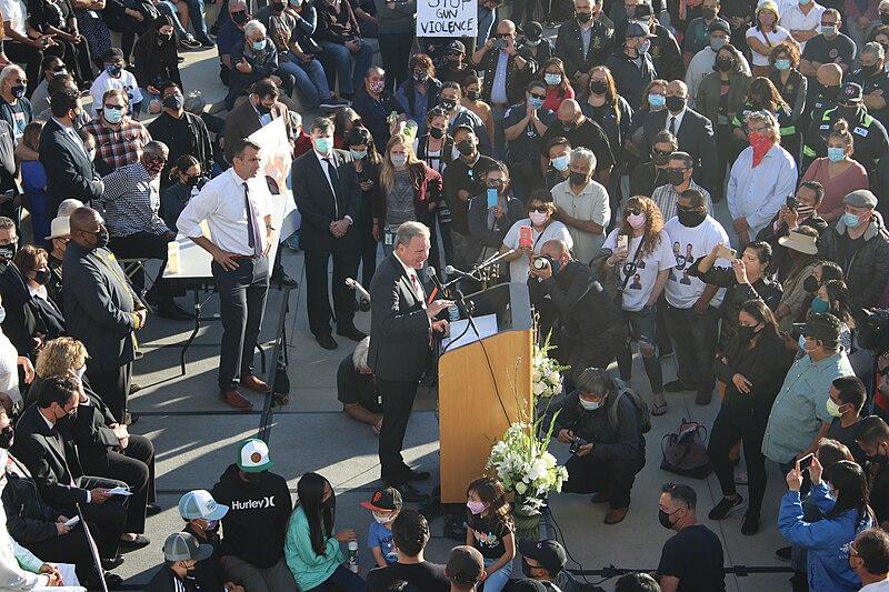 A masked crowd of hundreds surrounds an elected official giving a speech at a City Hall vigil for the victims of a 2021 shooting in San Jose at the Valley Transportation Agency railyards.