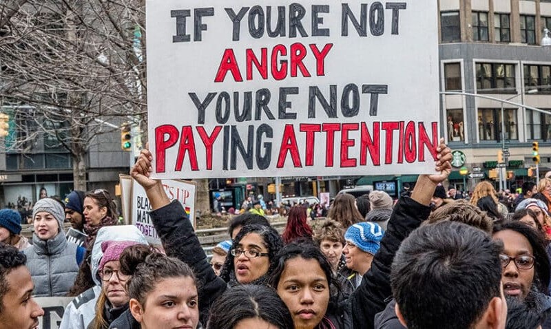 A photo of workers at a protest. The focus is on one's sign, which reads "If you're not angry, you're not paying attention"