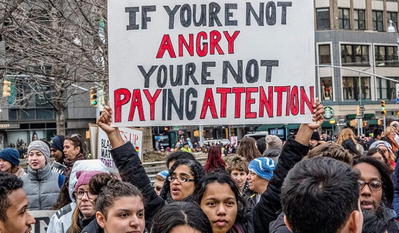 A photo of workers at a protest. One holds up a sign that says "If you're not angry, you're not paying attention"