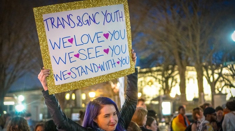 A white person with shoulder-length, side-parted purple hair holds up a sign in the middle of a dance protest at the White House. The sign reads Trans & GNC Youth, We Love You, We See You, We Stand With You.