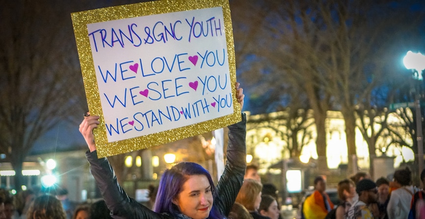 A white person with shoulder-length, side-parted purple hair holds up a sign in the middle of a dance protest at the White House. The sign reads Trans & GNC Youth, We Love You, We See You, We Stand With You.