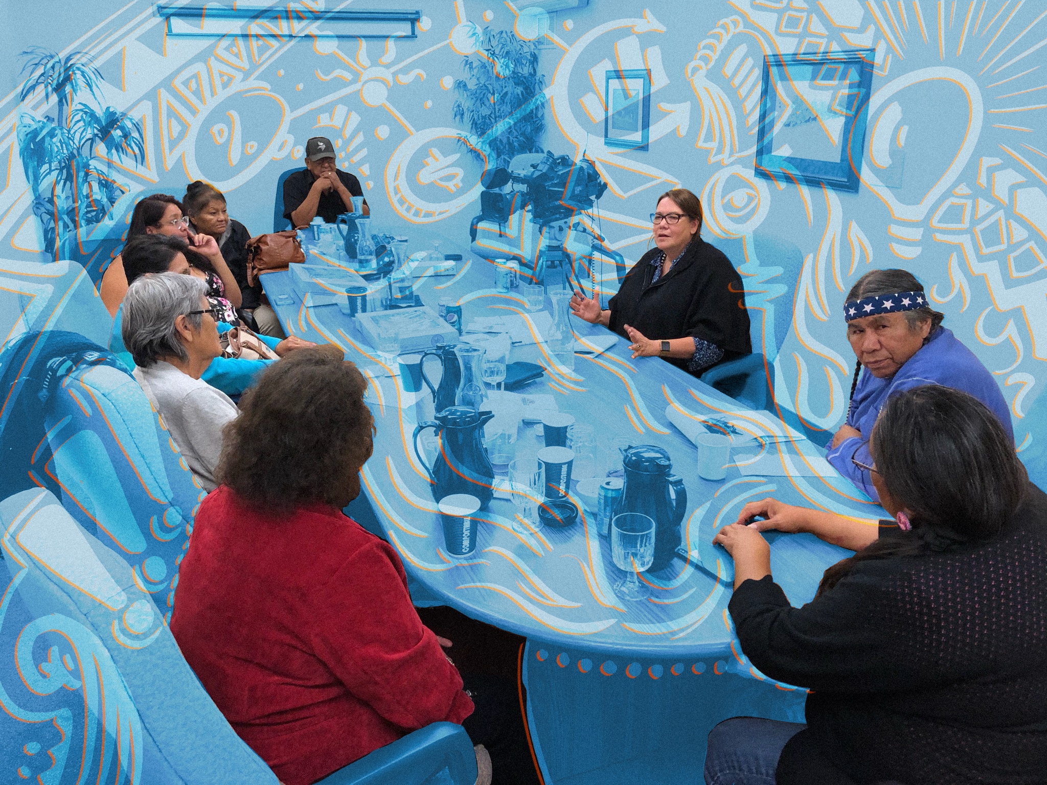 Nine Indigenous media-makers involved in the Indigenous Media Freedom Alliance are gathered around a table and talking. Jodi Rave Spotted Bear, the author, is sitting at upper middle-right, hands out for emphasis. There is a transparent blue overlay with decorative orange elements on the photo.