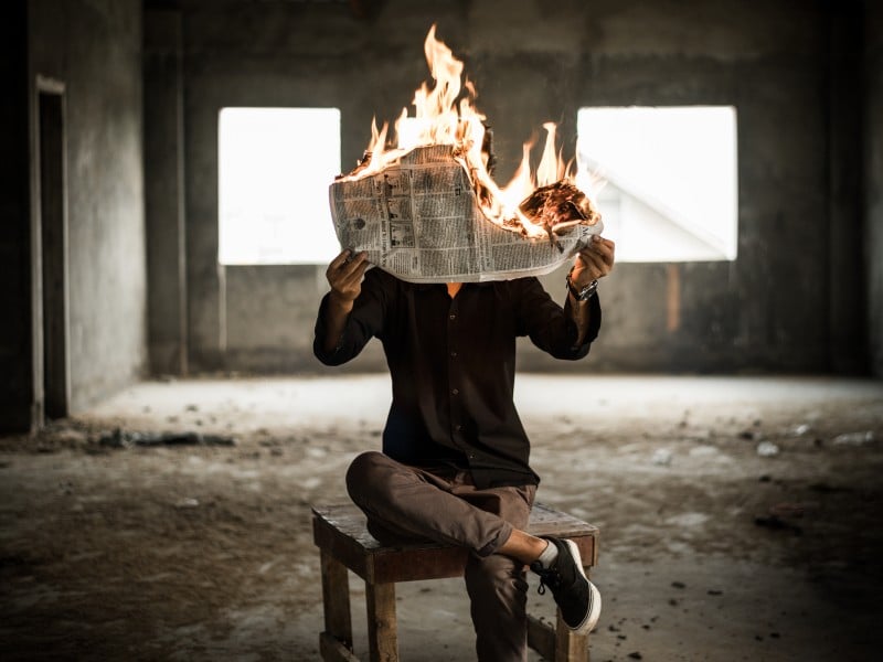 A person in a black button-down and brown pants sits on a stool holding up a burning newspaper in an empty room.