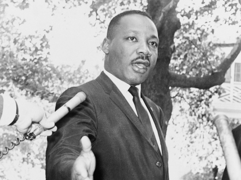 A black and white photo of Rev. Martin Luther King Jr. speaking to two microphones pointed at his face.