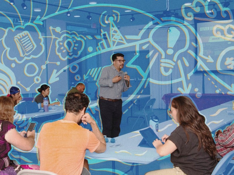 Anthony Victoria, a Latino man wearing a blue button-down and black pants, is holding a microphone and teaching at the center of a filled classroom. A blue transparent overlay is on the image with orange accents around the crowd and Anthony.