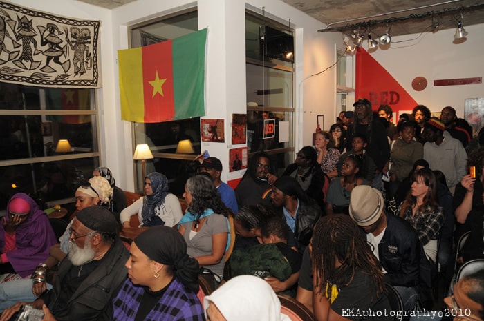 A majority Black crowd gathers at the Black Dot cafe. The interior is decorated with several posters, a tapestry of dancers, and the Cameroon flag.