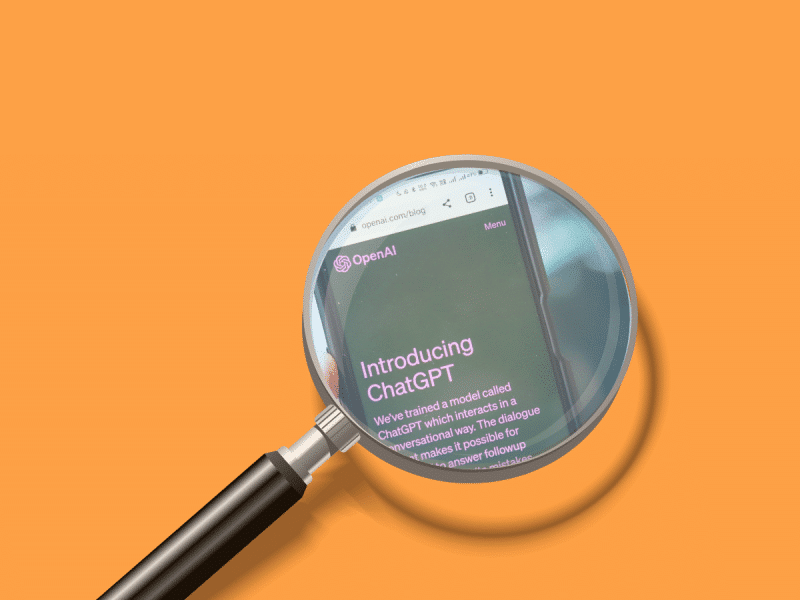On an orange background, a magnifying glass focused on a phone screen open to the home screen of ChatGPT through OpenAI's website.