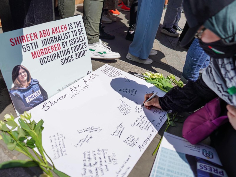 At a London vigil on May 14, 2022, an attendee wearing a mask with the Palestine flag and a keffiyeh kneels and writes condolences on a poster dedicated to Shireen Abu Akleh. Propped up in front of that poster is another poster with a photo of Shireen in a press vest, reading "Shireen Abu Akleh is the 55th journalist to be murdered by Israeli occupation forces since 2000." That statistic is cited from the Palestinian Journalists Syndicate.