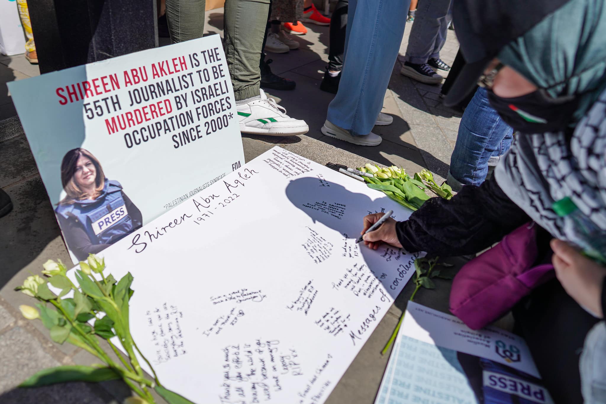 At a London vigil on May 14, 2022, an attendee wearing a mask with the Palestine flag and a keffiyeh kneels and writes condolences on a poster dedicated to Shireen Abu Akleh. Propped up in front of that poster is another poster with a photo of Shireen in a press vest, reading "Shireen Abu Akleh is the 55th journalist to be murdered by Israeli occupation forces since 2000." That statistic is cited from the Palestinian Journalists Syndicate.

 