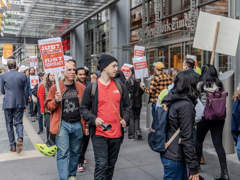 New York Times Tech Guild members and their supporters walk the picket line in front of The New York Times office building holding signs that say "Times Tech Guild: Just practicing for a Just Contract" and "Times Tech Guild: On ULP Strike."