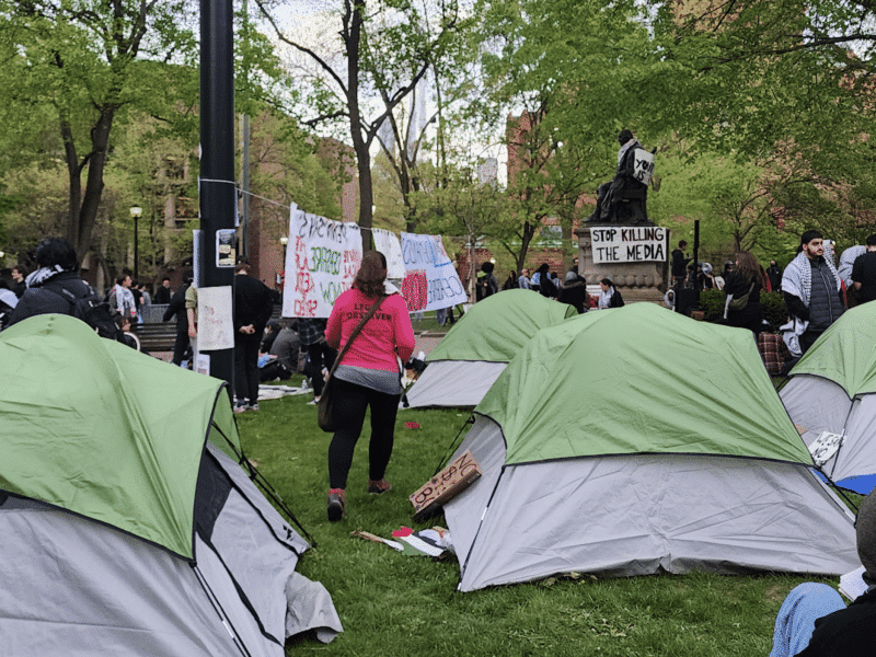 A legal observer walks between tents at the University of Pennsylvania's Palestine solidarity encampment. A poster displayed prominently hung on a statue says "Stop Killing the Media."