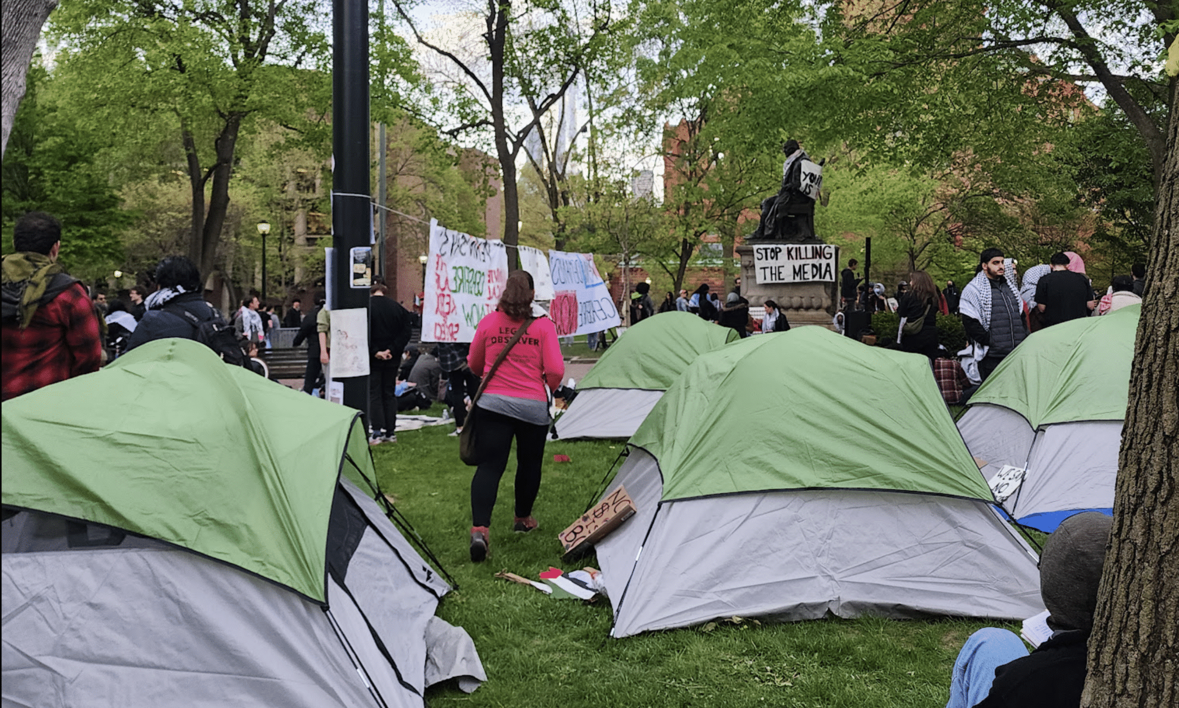 A legal observer walks between tents at the University of Pennsylvania's Palestine solidarity encampment. A poster displayed prominently hung on a statue says "Stop Killing the Media." 