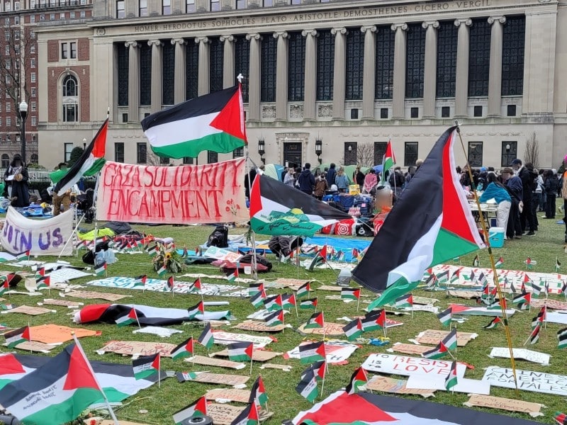 Palestine flags sit among tents at the Gaza Solidarity Encampment on a lawn on Columbia University as people, faces blurred, mill about behind the encampment's entrance.