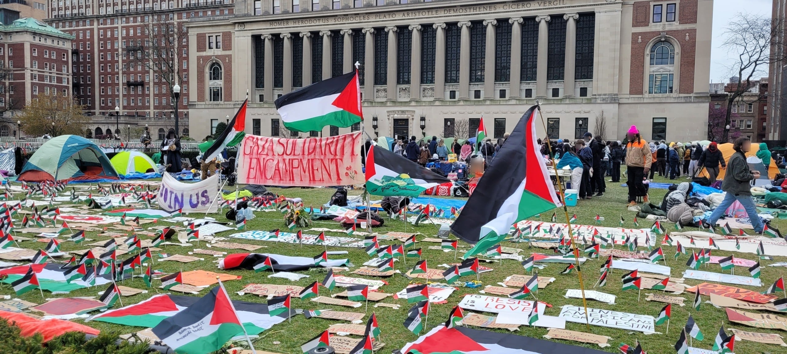 Palestine flags sit among tents at the Gaza Solidarity Encampment on a lawn on Columbia University as people, faces blurred, mill about behind the encampment's entrance.