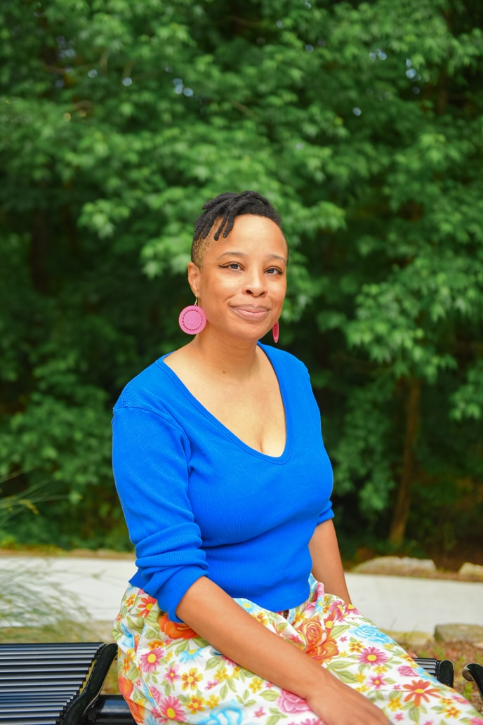 A smiling Black person with short natural black hair wears a blue shirt, flowered pants, and pink earrings while sitting on a bench at Cascade Springs Nature Preserve, Atlanta, Georgia, USA. 