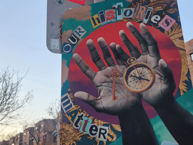 A mural on the back of a lamppost. Cut-out letters read Our Histories Matter around a red and purple circle behind a photo of Black hands holding a key and a compass.