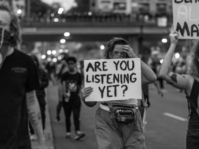 A grayscale photo of a group of masked protesters holding up signs; the foremost says "Are You Listening Yet?"