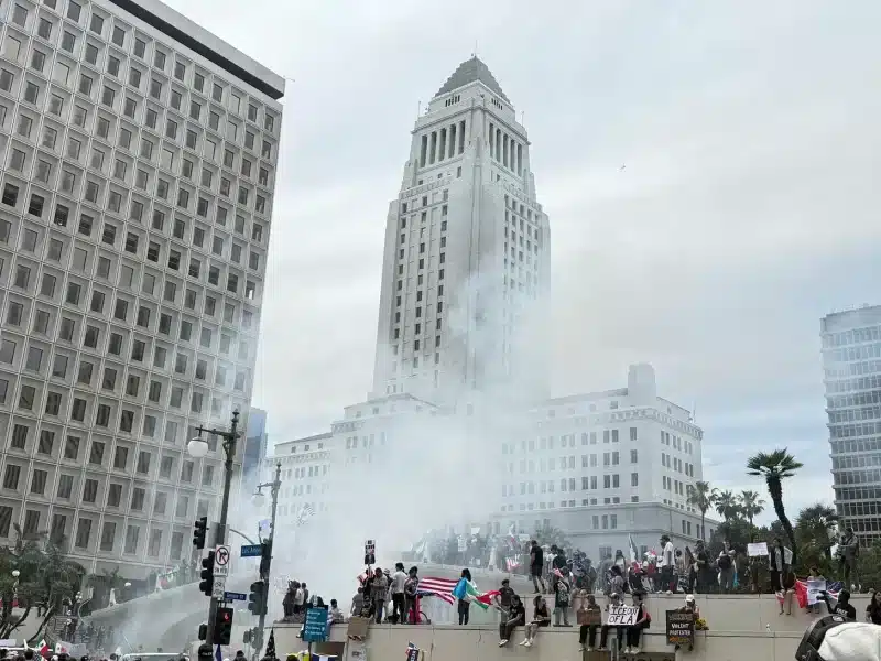 A crowd gathers around Los Angeles City Hall as smoke rises from the tires of a driver performing donuts in front of the building.