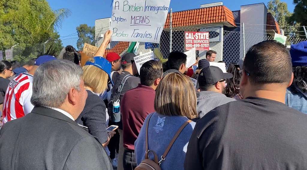 A crowd of people gathers to protest outside a new ICE detention facility in Arleta. One person holds a poster saying "A broken immigration system means broken families."
