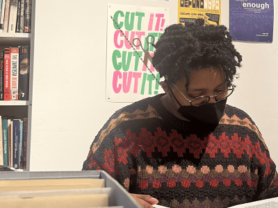 A masked Black person with short twists and glasses pores over records at a library.