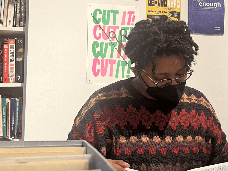A masked Black person with short twists and glasses pores over records at a library.