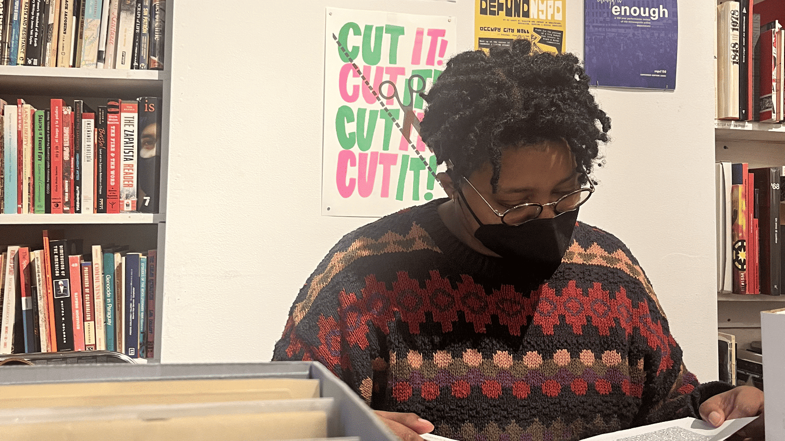 A masked Black person with short twists and glasses pores over records at a library. 