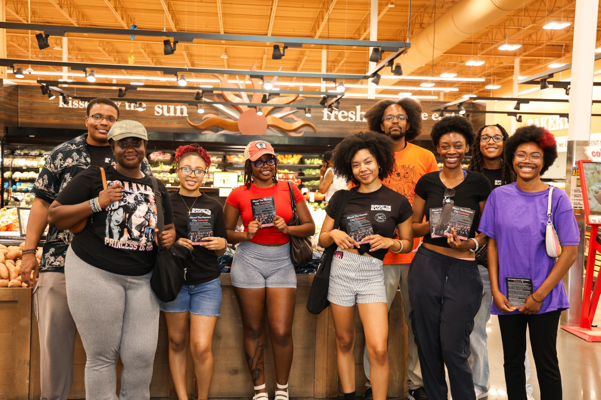 Nine young Black people pose in an otherwise empty produce section of a grocery store, holding up flyers about the KC Defender.