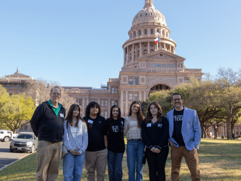 Seven people — two advisors and five high schoolers — stand in front of the Texas Capitol building.